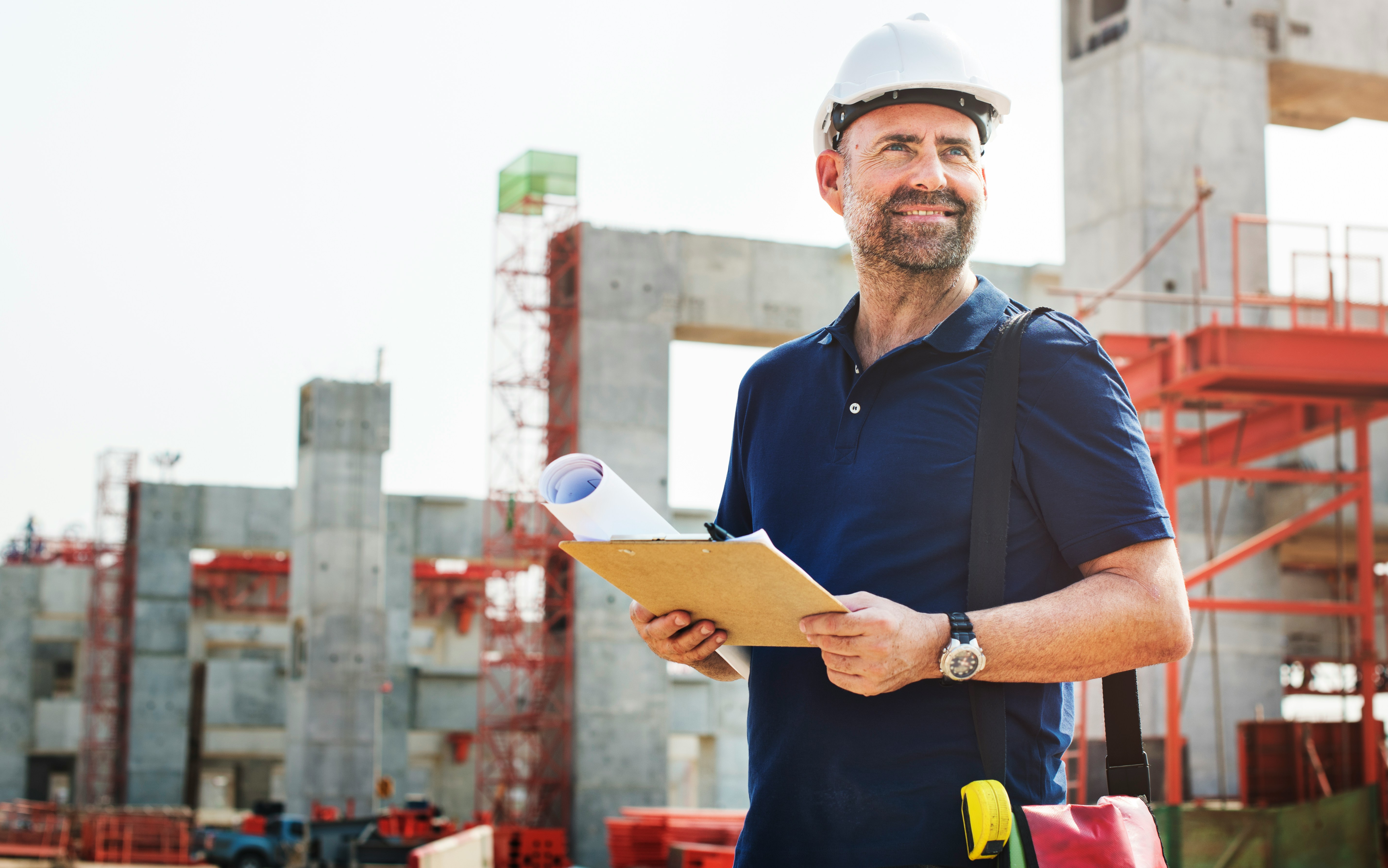 Construction worker wearing a white hard hat and navy blue shirt holding blueprints and clipboard at a building site.