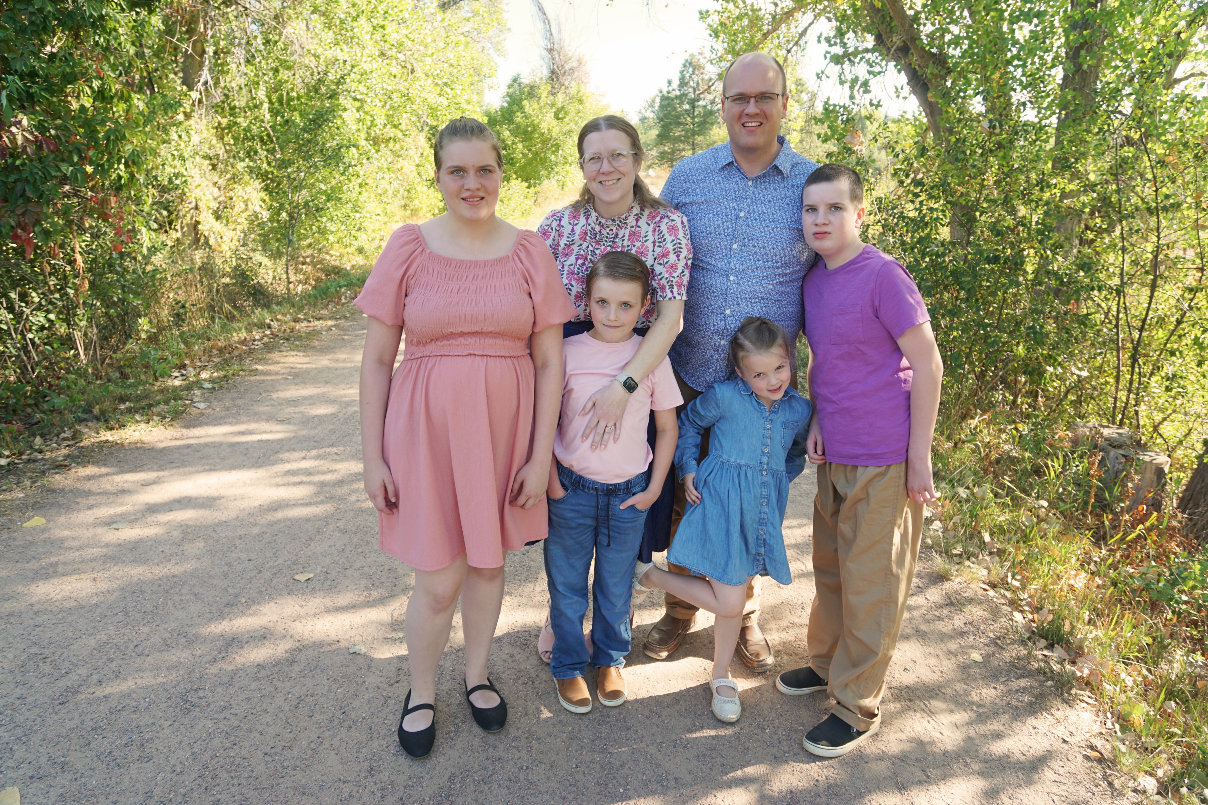 The Johnson family, A family of six standing together on a sunlit path surrounded by green trees, smiling at the camera.