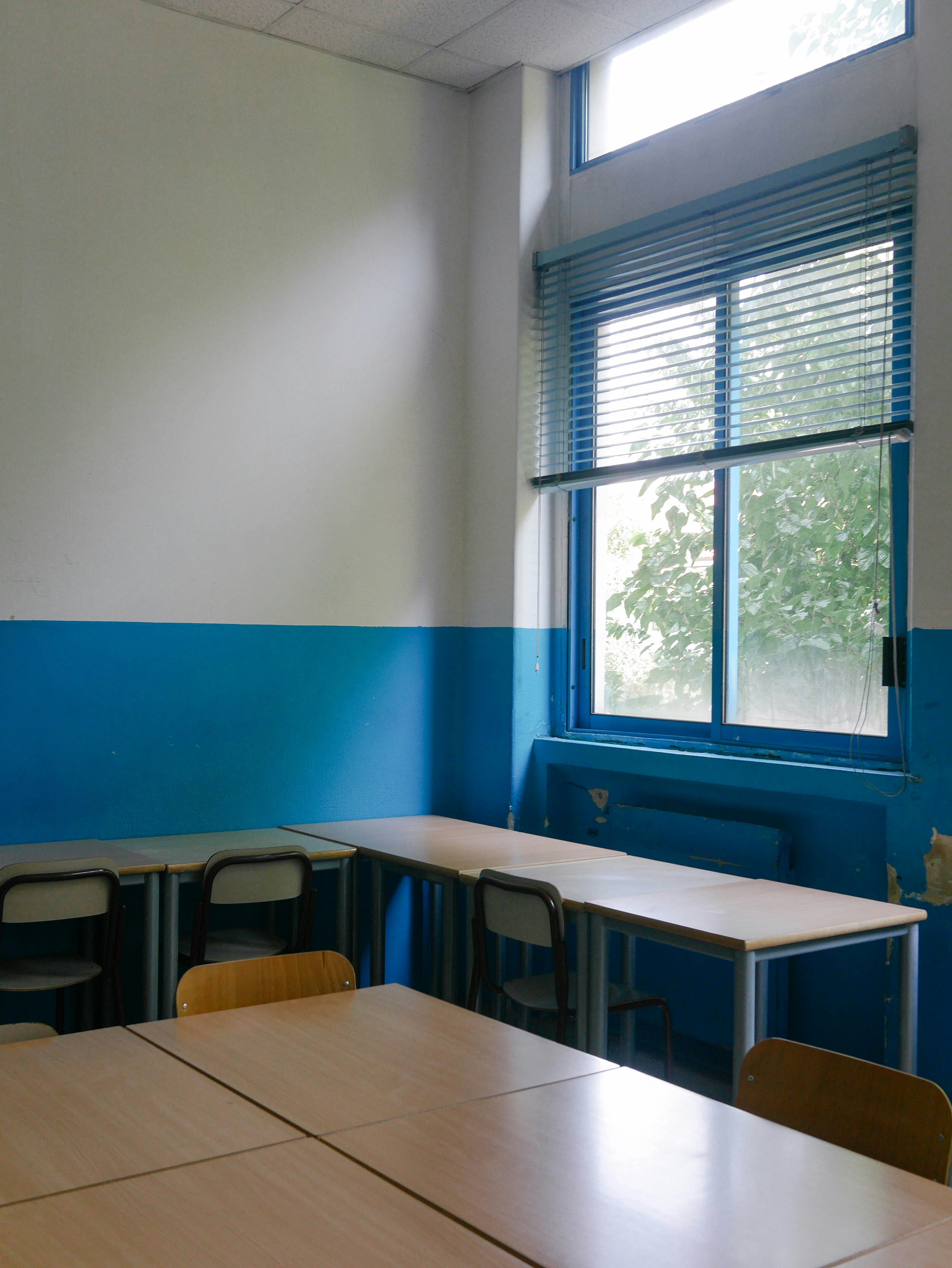 Bright classroom with blue and white walls, wooden tables, black chairs, and large window with blue blinds.