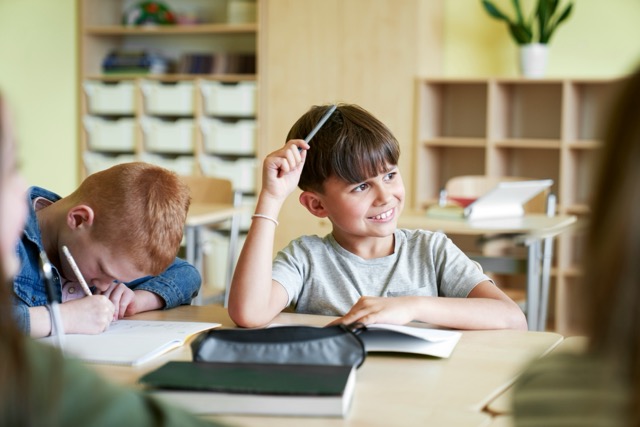 Two students sit at desk in classroom, one reads while other scratches head thoughtfully.