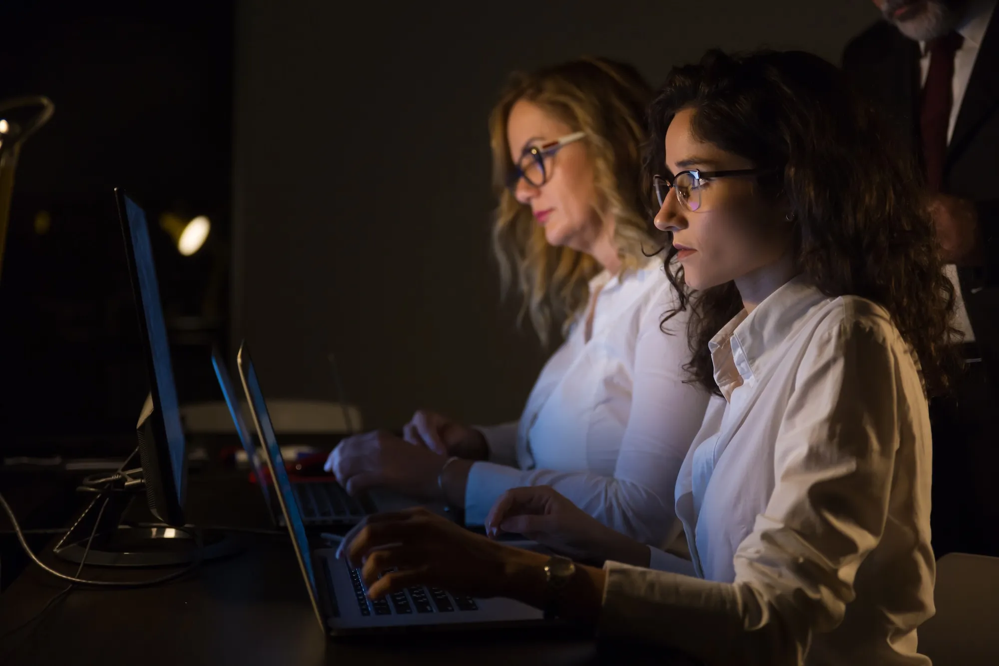 Two serious women wearing glasses working on laptops in a dimly lit room.
