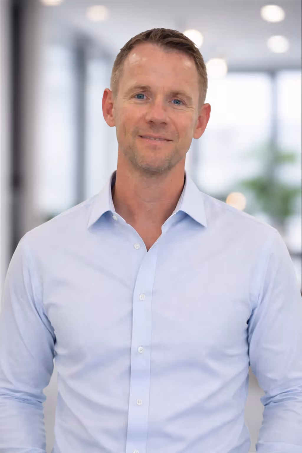 Smiling man in a light blue buttoned shirt standing in a bright office with blurred background.