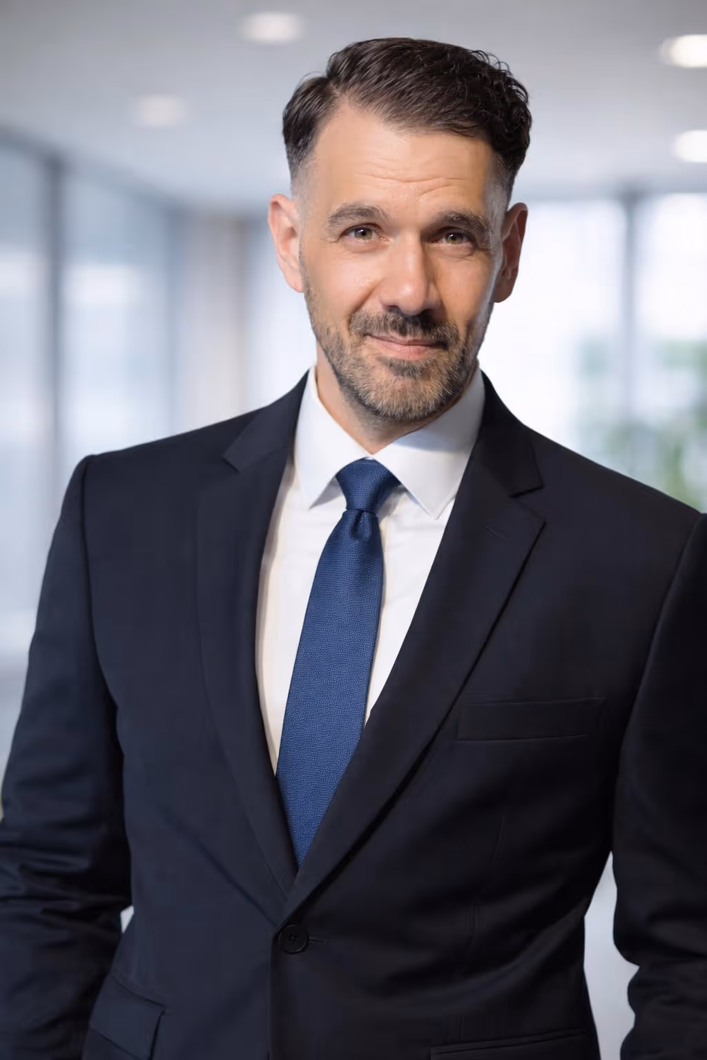 Confident middle-aged man in a dark suit and blue tie smiling in a bright office setting.