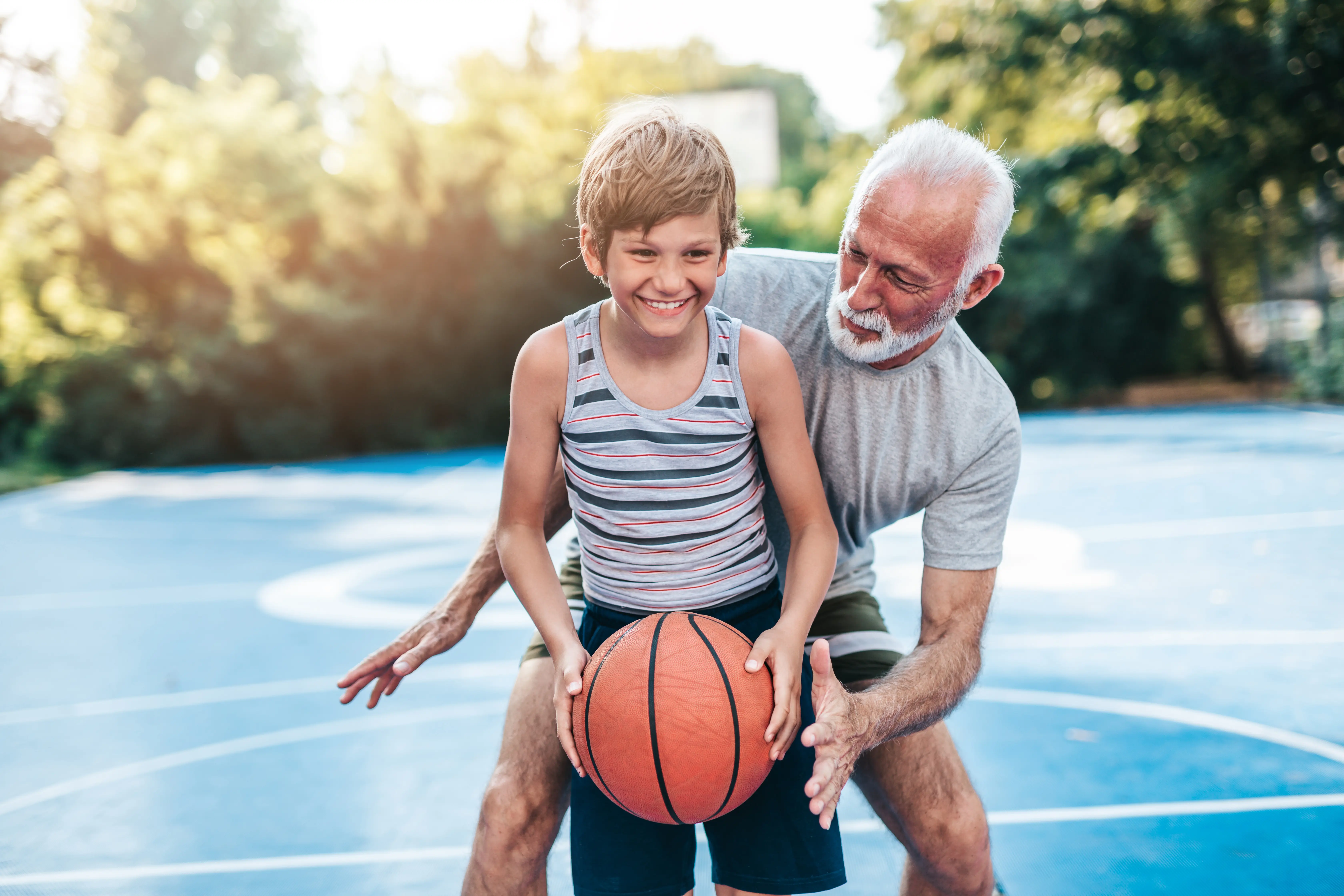 An active elderly man, able to participate in sports likely due to effective chiropractic care, teaches a young boy how to hold a basketball on an outdoor court, both sharing a joyful moment.