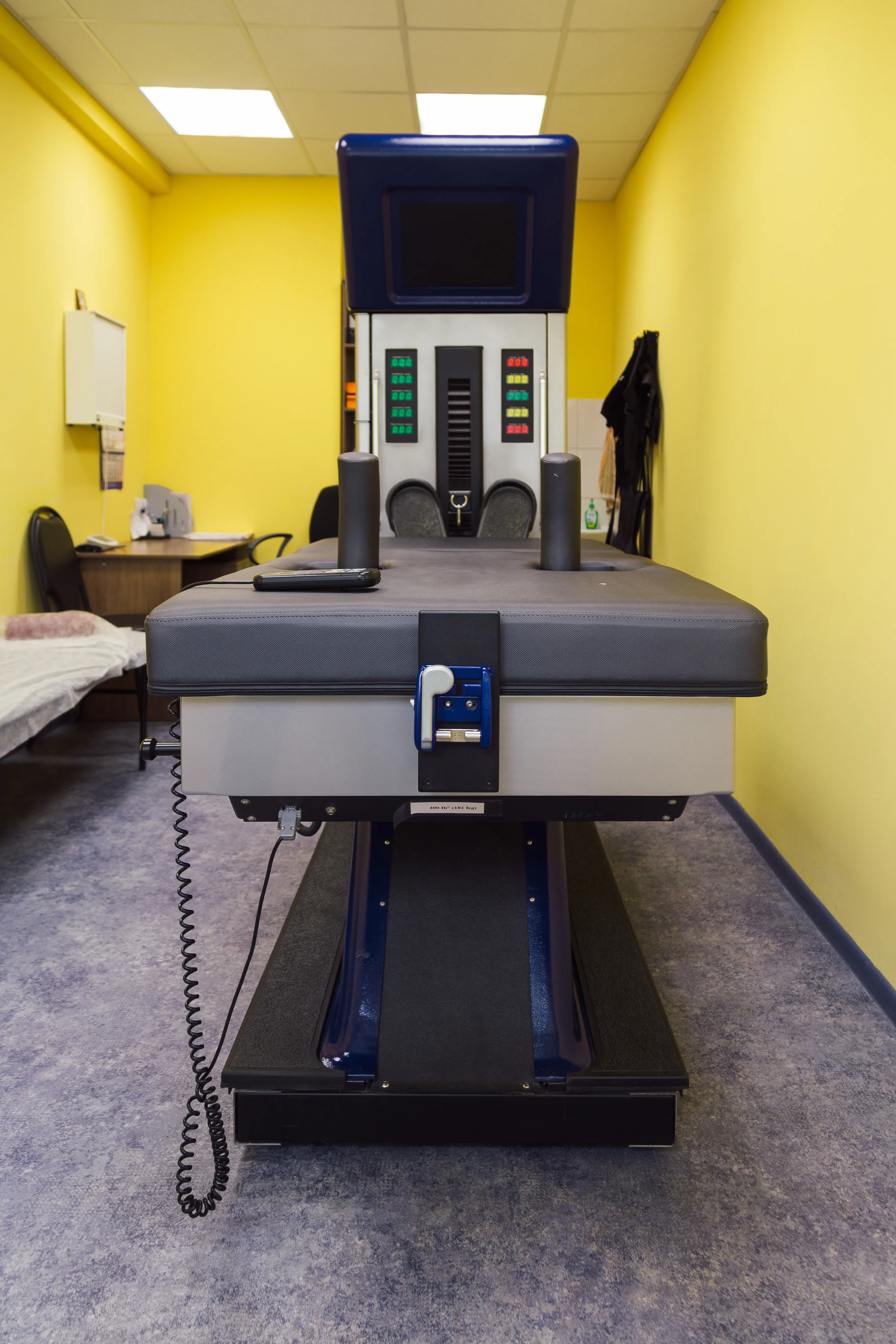 An advanced chiropractic treatment table featuring electronic controls and a screen, situated in a yellow-walled room with a clinical setup.