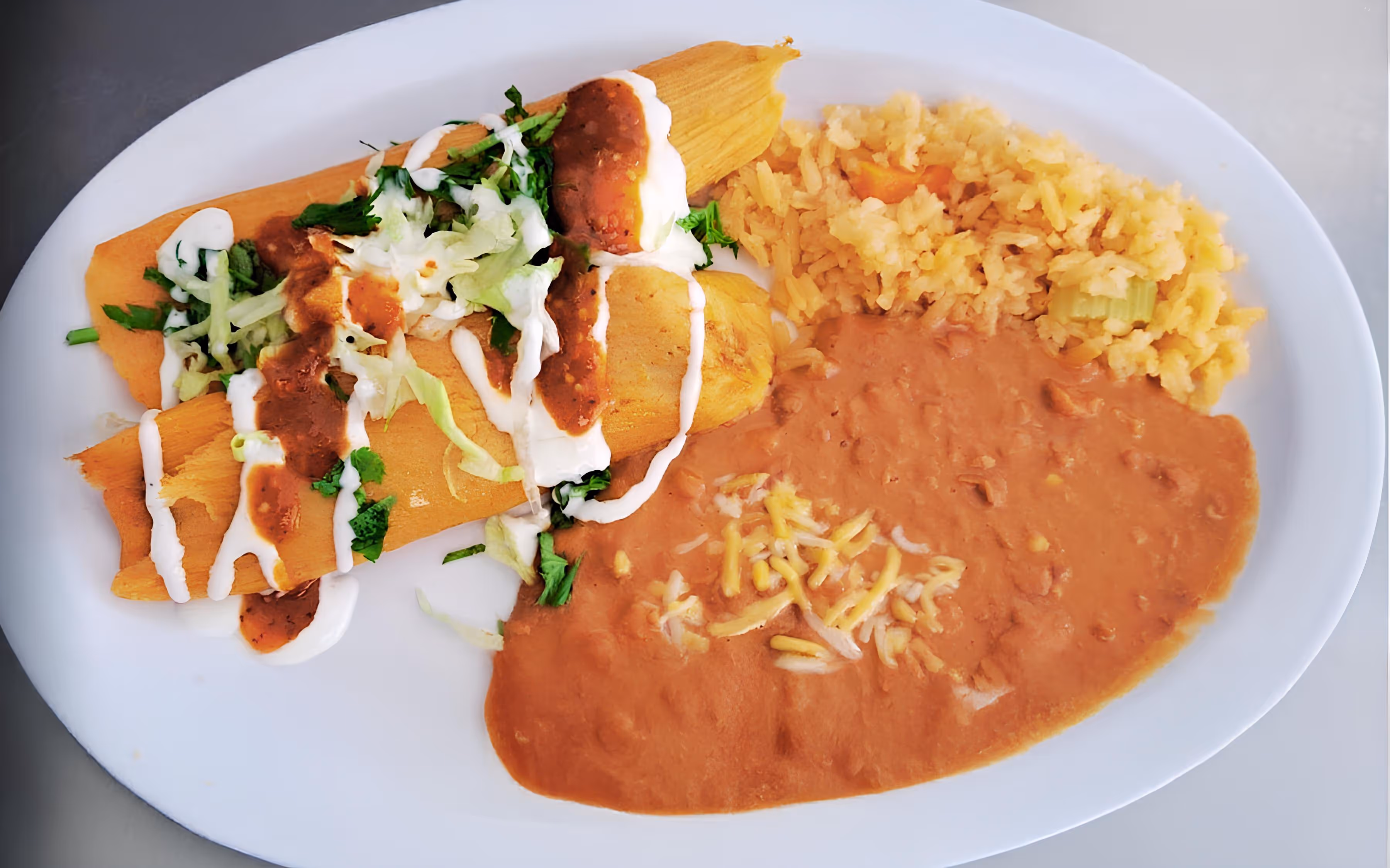 A plate with two tamales topped with sour cream, red salsa, and lettuce, accompanied by refried beans garnished with cheese and a serving of Mexican rice.