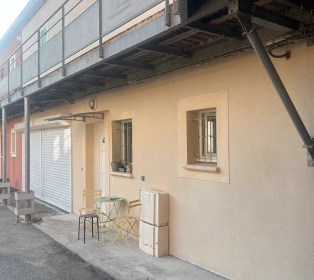 Façade rose pâle d'une maison avec un garage blanc, trois fenêtres dont une avec volets fermés, et une unité de climatisation extérieure.