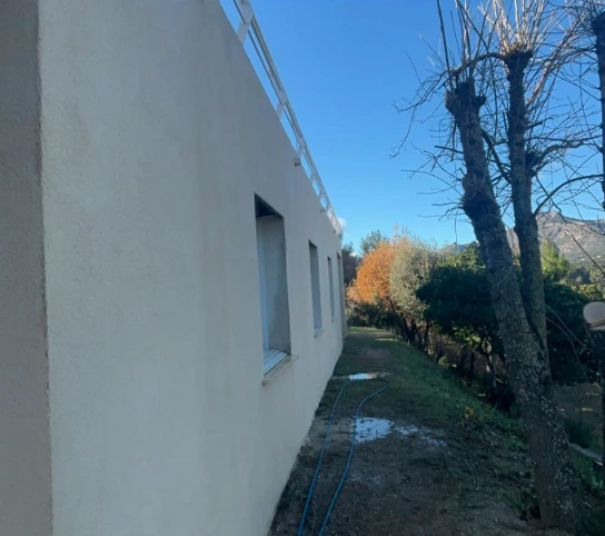 Façade jaune d'une maison avec volets blancs fermés, ombragée par un arbre au feuillage vert sous un ciel bleu avec quelques nuages.