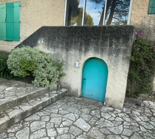 Façade extérieure d'une maison avec volets blancs ouverts, un mur en béton et des arbres verts sous un ciel bleu clair.