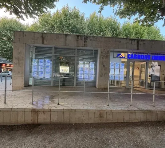 Façade extérieure d'une maison avec volets blancs ouverts, un mur en béton et des arbres verts sous un ciel bleu clair.