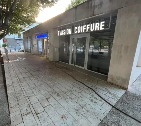 Façade extérieure d'une maison avec volets blancs ouverts, un mur en béton et des arbres verts sous un ciel bleu clair.