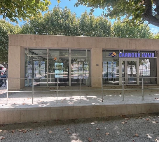 Façade jaune d'une maison avec volets blancs fermés, ombragée par un arbre au feuillage vert sous un ciel bleu avec quelques nuages.