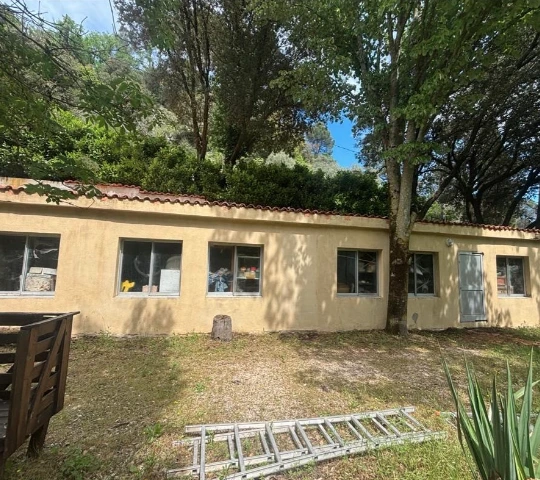 Façade jaune d'une maison avec volets blancs fermés, ombragée par un arbre au feuillage vert sous un ciel bleu avec quelques nuages.