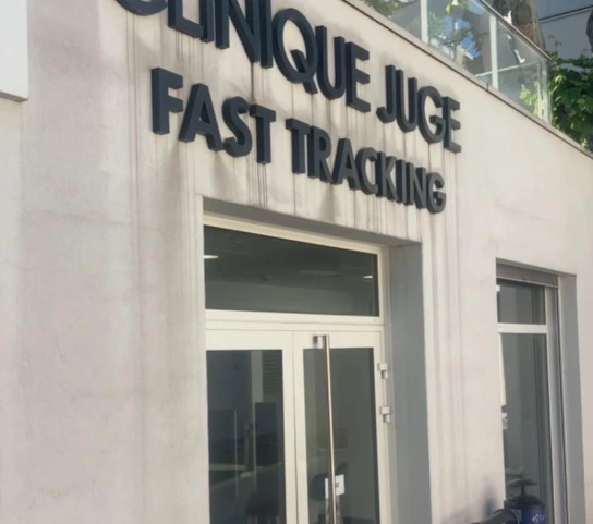 Façade extérieure d'une maison avec volets blancs ouverts, un mur en béton et des arbres verts sous un ciel bleu clair.