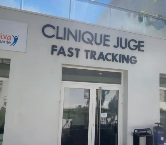 Façade jaune d'une maison avec volets blancs fermés, ombragée par un arbre au feuillage vert sous un ciel bleu avec quelques nuages.