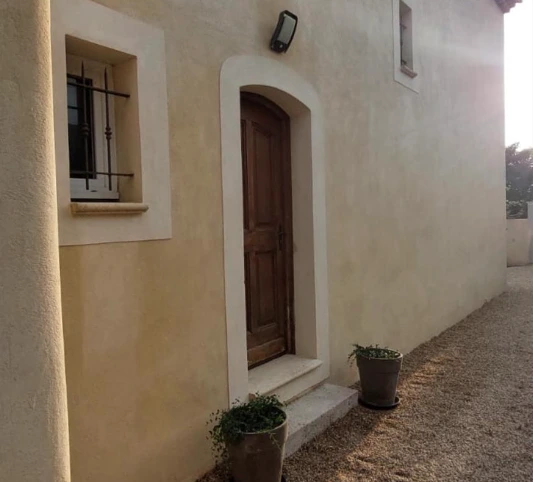 Façade jaune d'une maison avec volets blancs fermés, ombragée par un arbre au feuillage vert sous un ciel bleu avec quelques nuages.