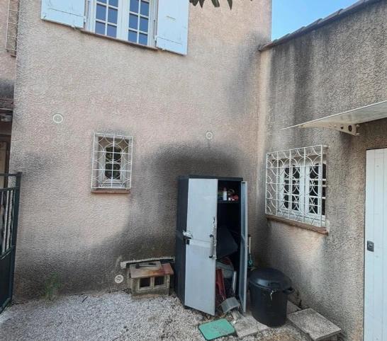 Façade extérieure d'une maison avec volets blancs ouverts, un mur en béton et des arbres verts sous un ciel bleu clair.