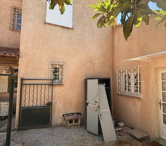 Façade jaune d'une maison avec volets blancs fermés, ombragée par un arbre au feuillage vert sous un ciel bleu avec quelques nuages.