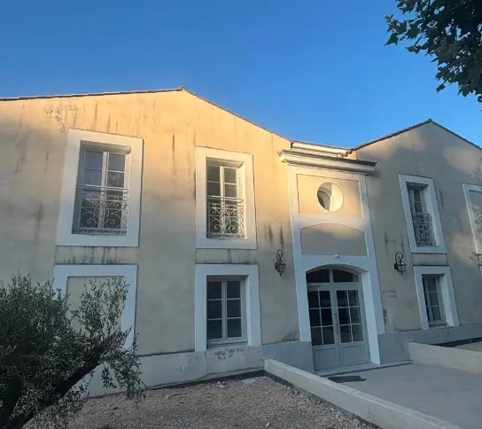 Façade extérieure d'une maison avec volets blancs ouverts, un mur en béton et des arbres verts sous un ciel bleu clair.