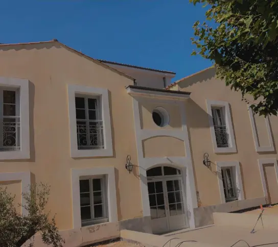 Façade jaune d'une maison avec volets blancs fermés, ombragée par un arbre au feuillage vert sous un ciel bleu avec quelques nuages.