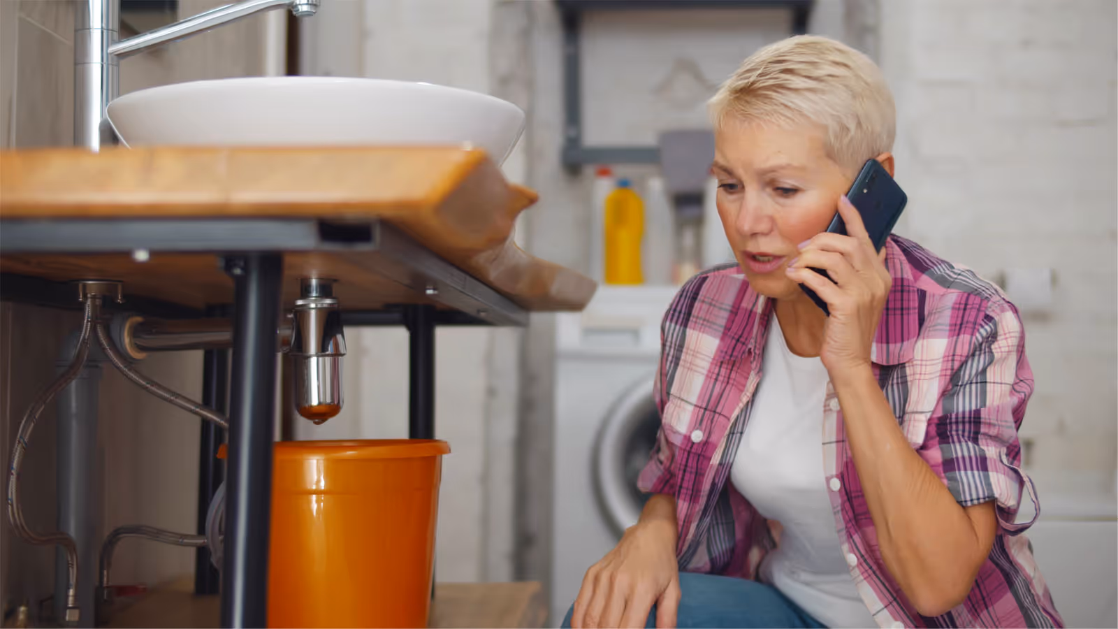Woman in a pink plaid shirt calling for plumbing help while a faucet leaks above an orange bucket.