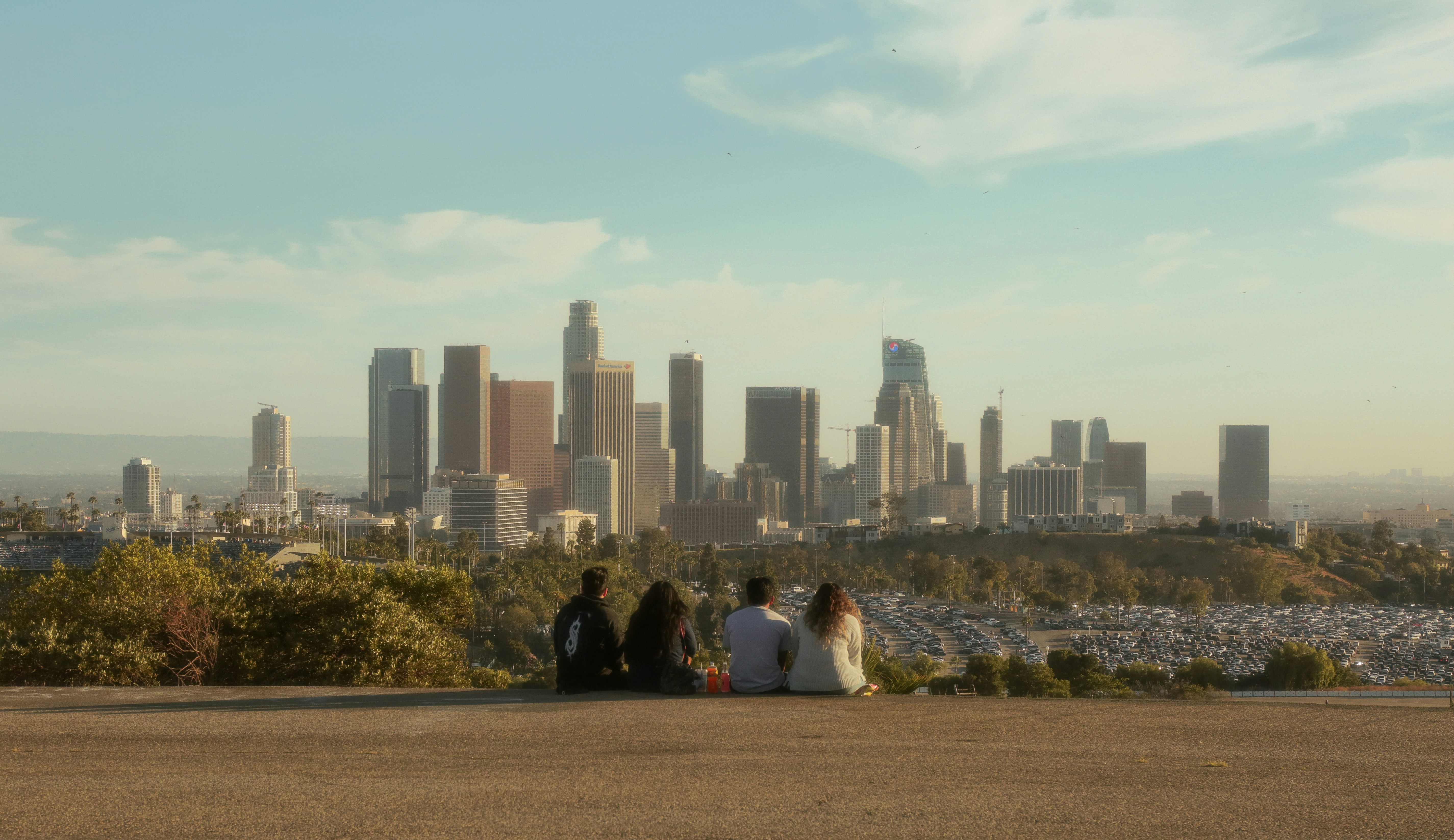 Four people sitting on a ledge overlooking downtown Los Angeles skyline during daylight.