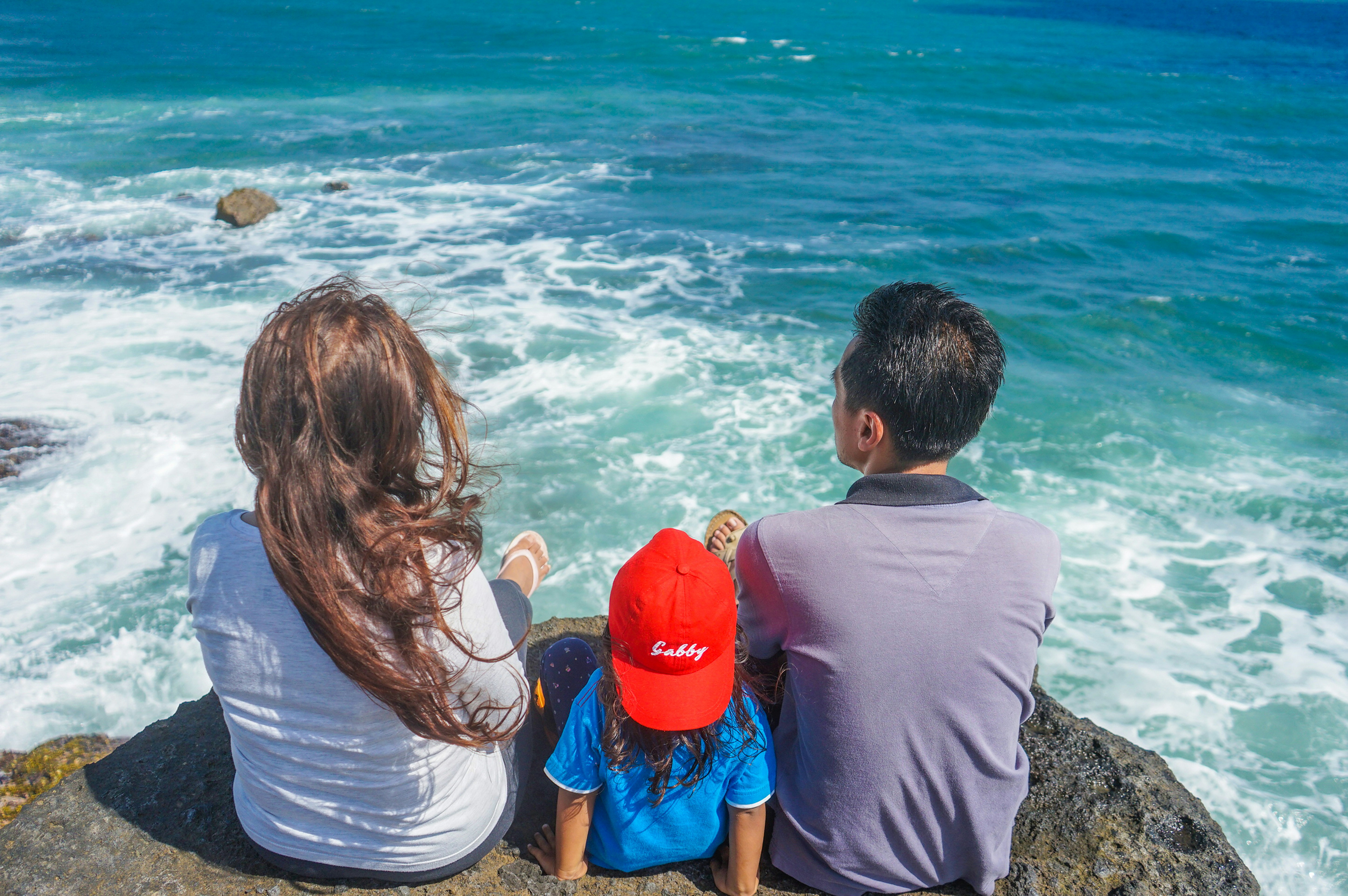 Family of three sitting on rocks overlooking the ocean with waves crashing below.
