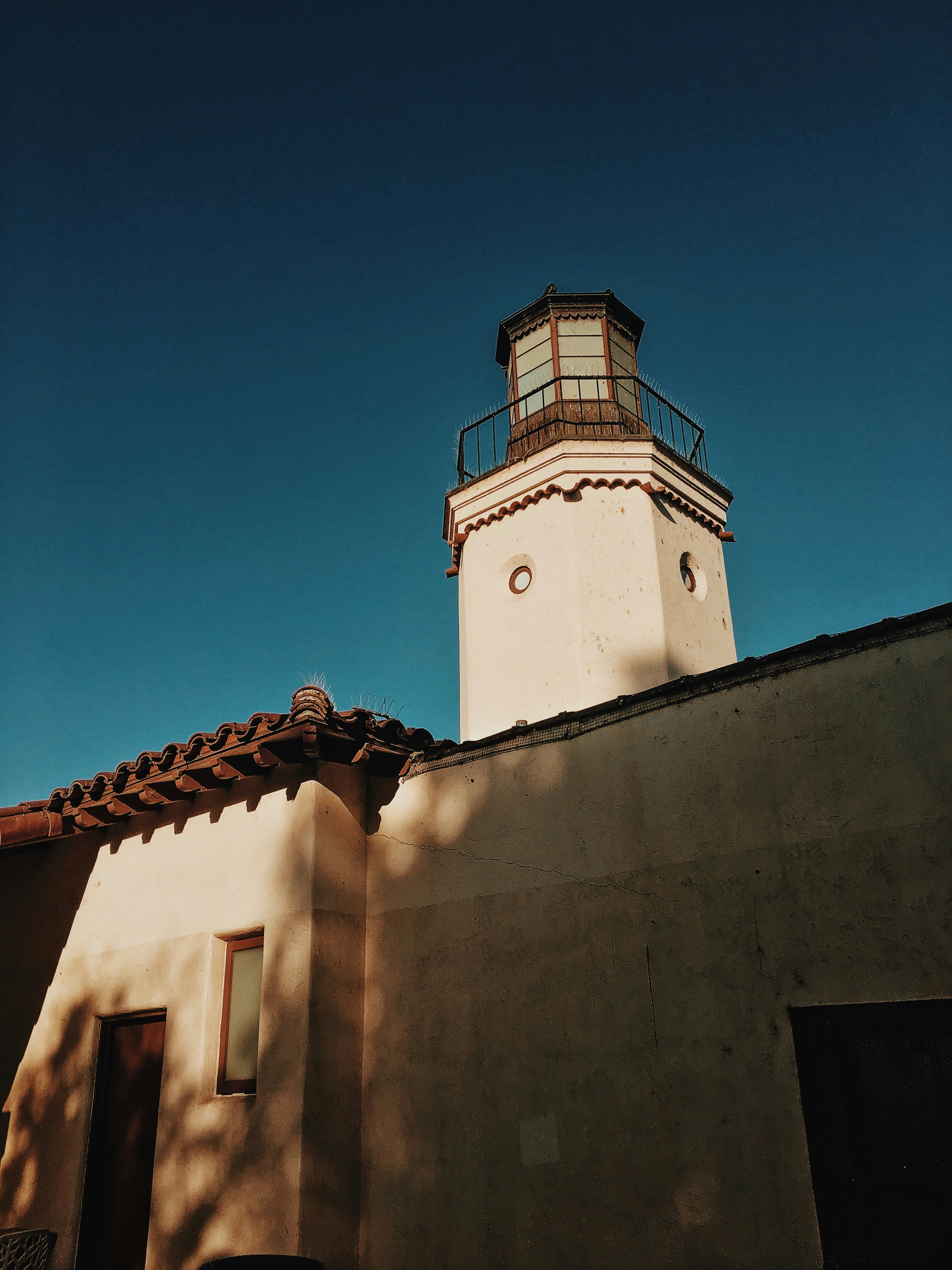 White lighthouse tower with a railing under a clear blue sky above a building with a tiled roof.