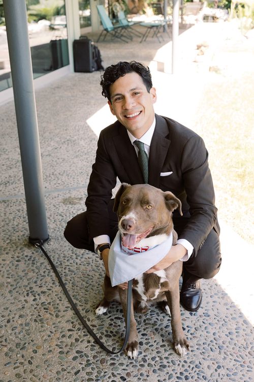 Man in a dark suit crouching and smiling beside a brown dog wearing a white bandana on a stone patio.