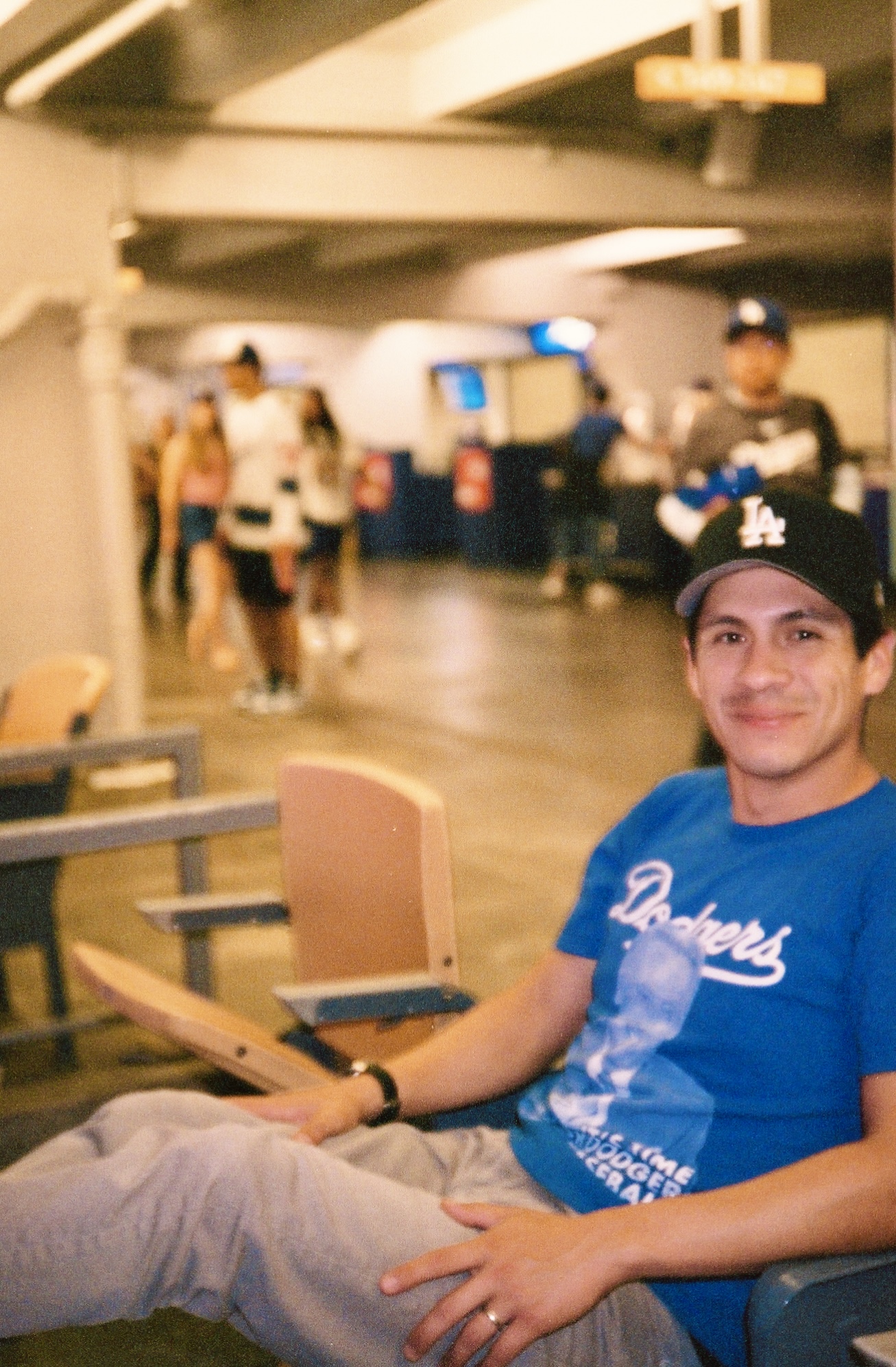 Man smiling and sitting in a stadium or arena seating area wearing a blue Dodgers shirt and black LA baseball cap.
