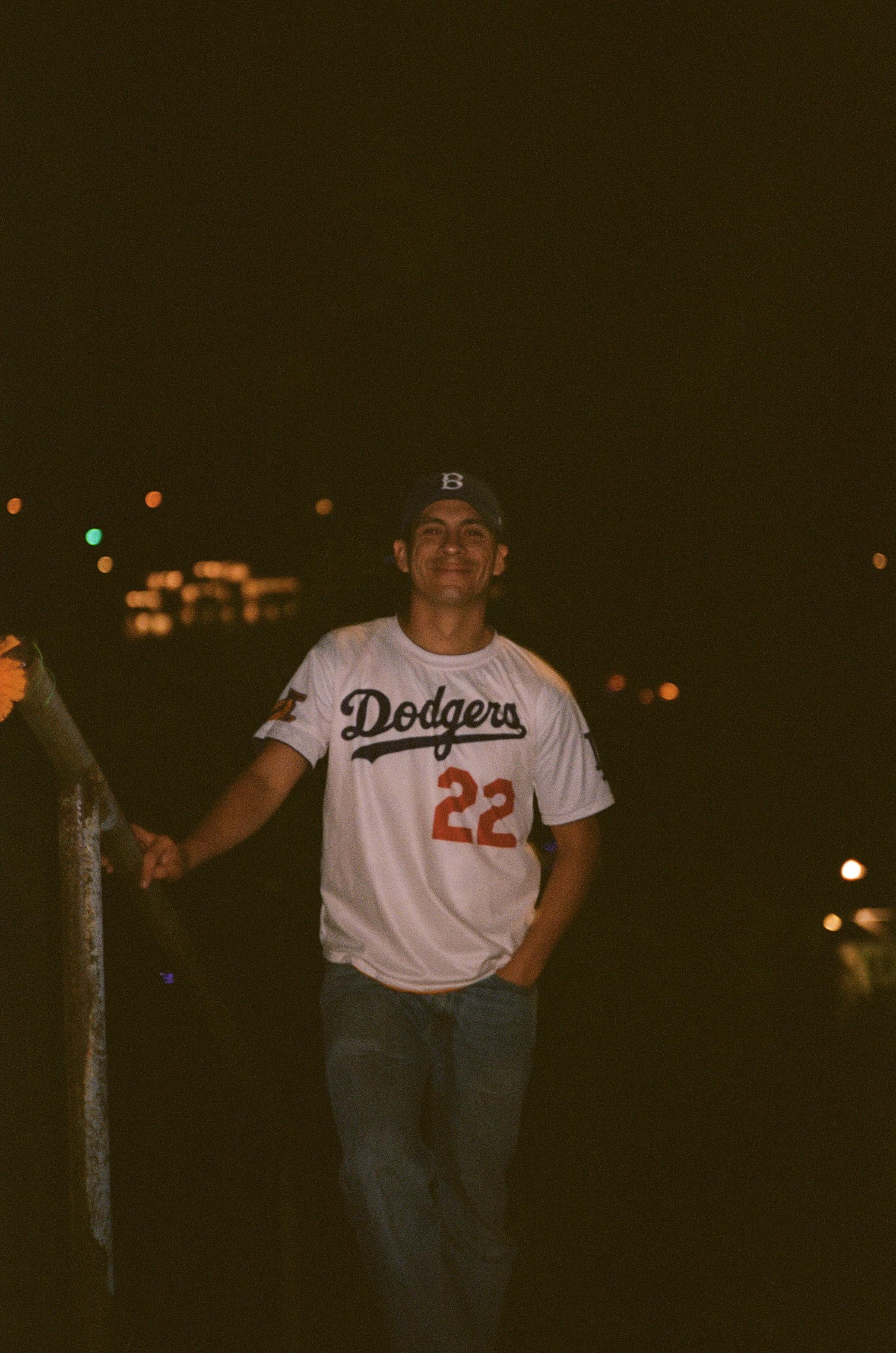 A man wearing a white Dodgers baseball jersey and a cap, standing outdoors at night with one hand on a railing and smiling.