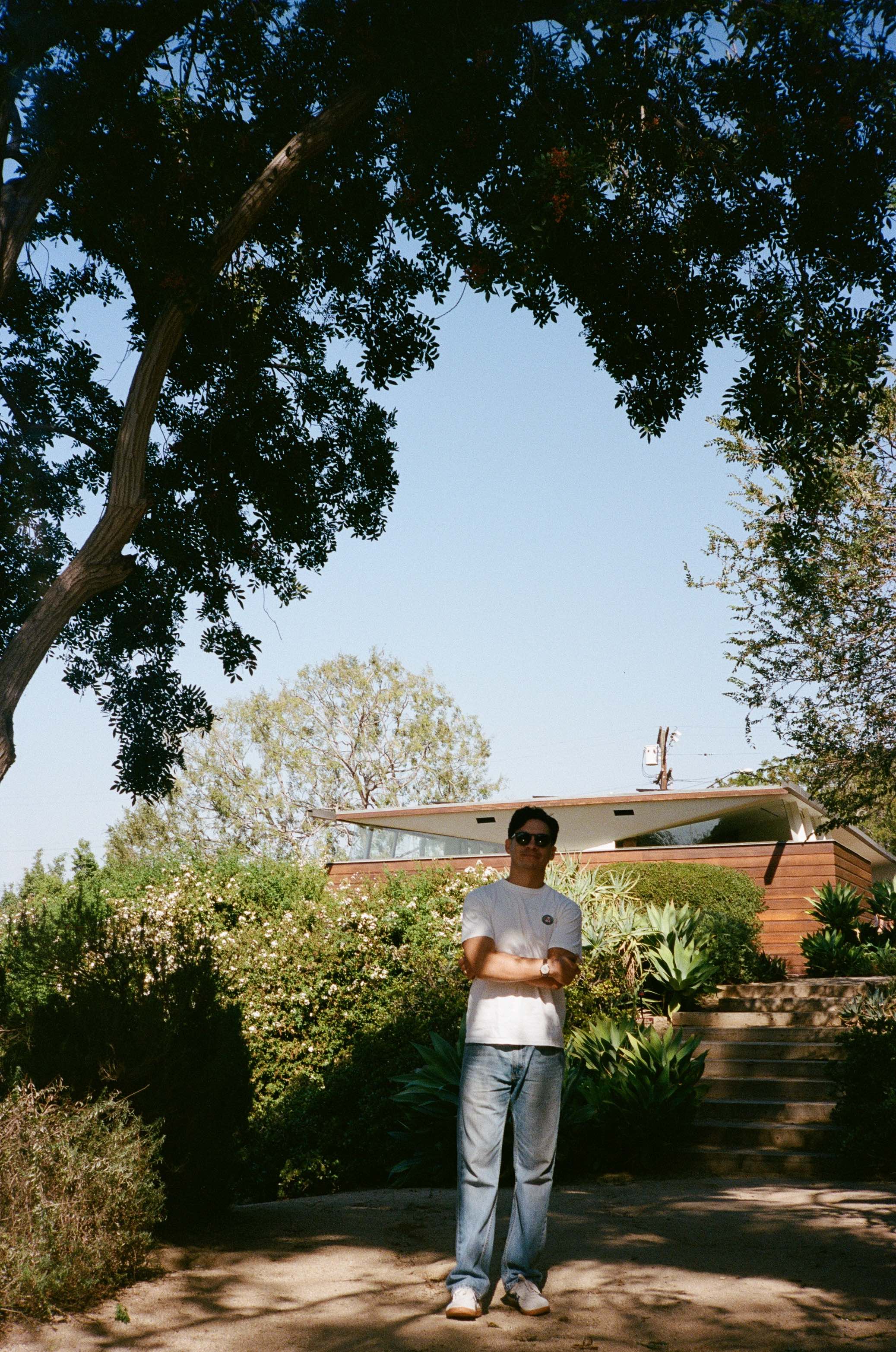 Man wearing sunglasses, white t-shirt, and jeans standing outdoors in front of a modern wooden house with greenery and trees around.