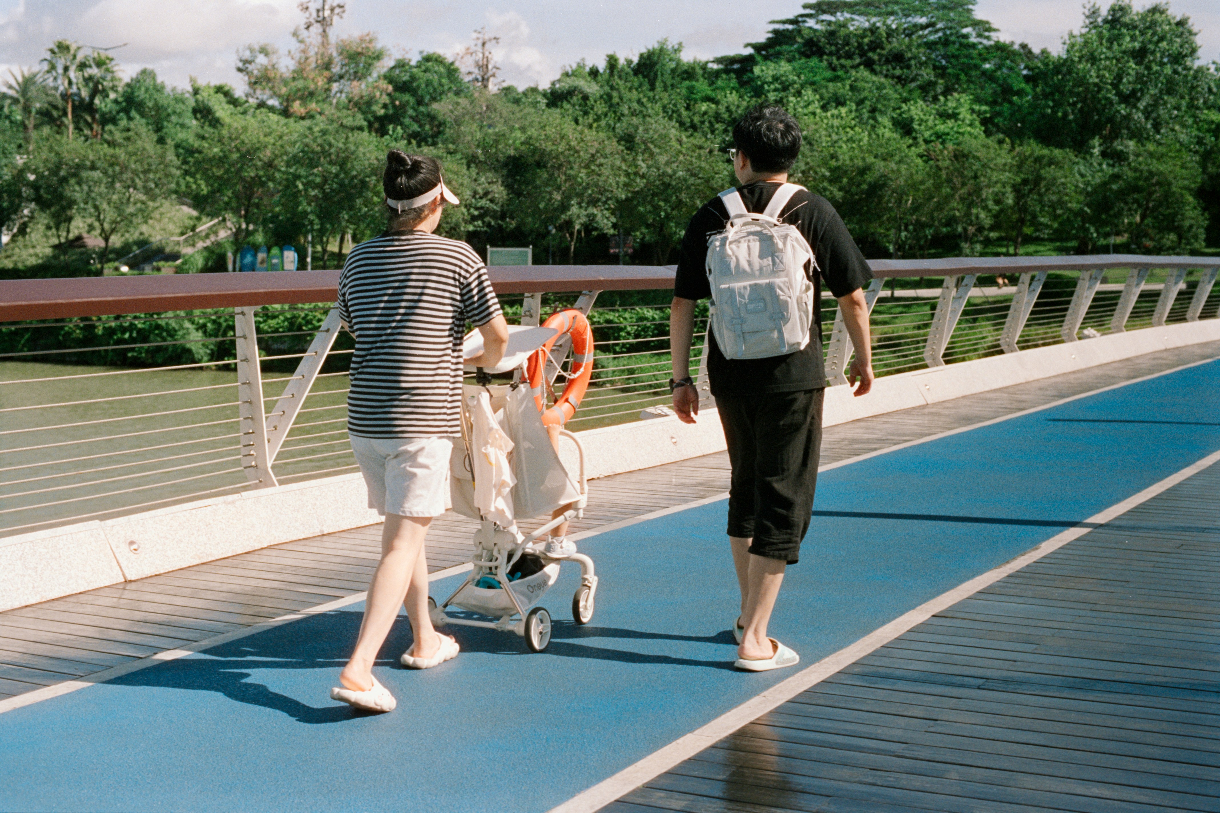 Two people walking on a blue pathway on a bridge, one pushing a stroller with a child inside.