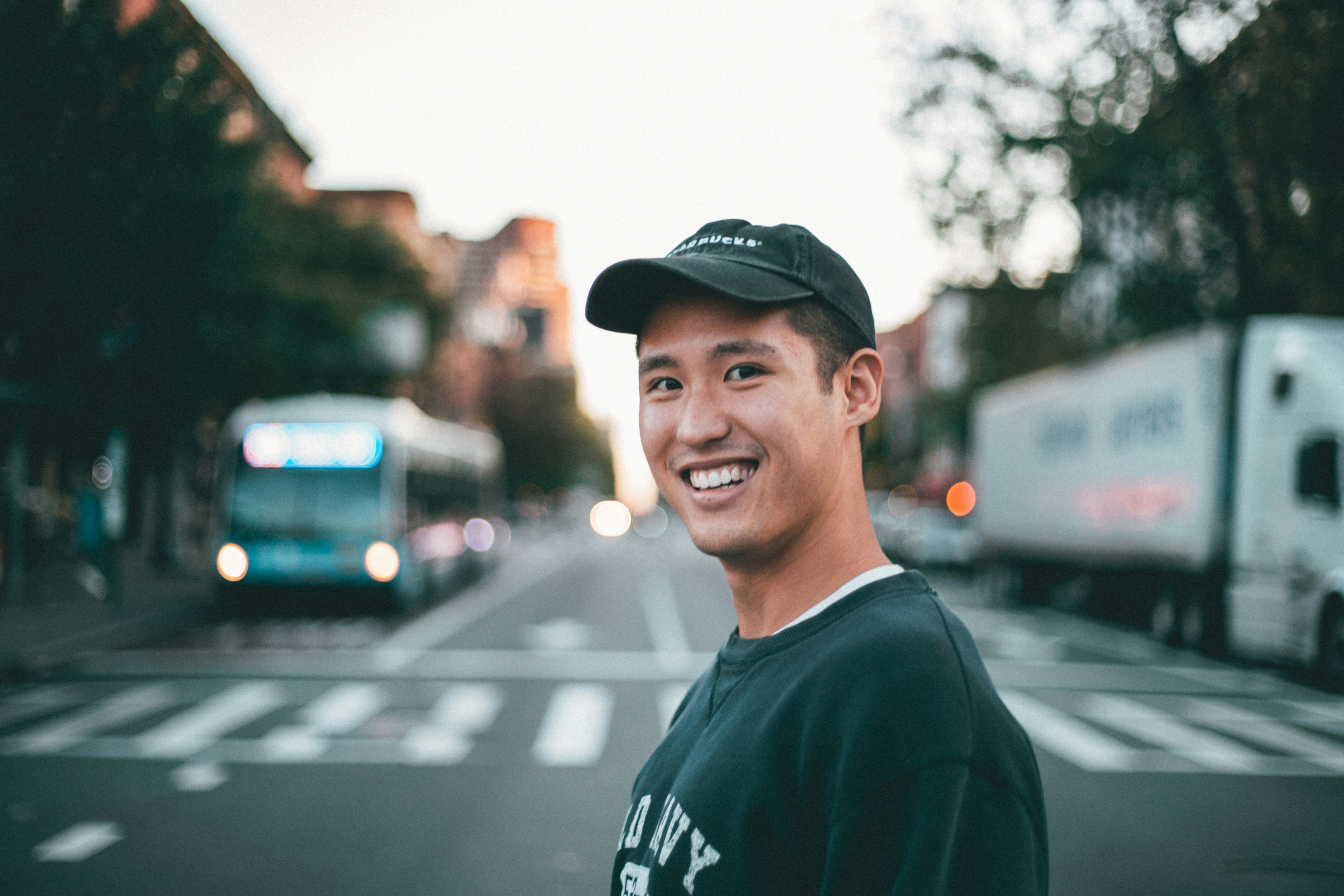 Smiling young man wearing a black cap and dark sweatshirt standing on a city street with blurred bus and truck in the background.