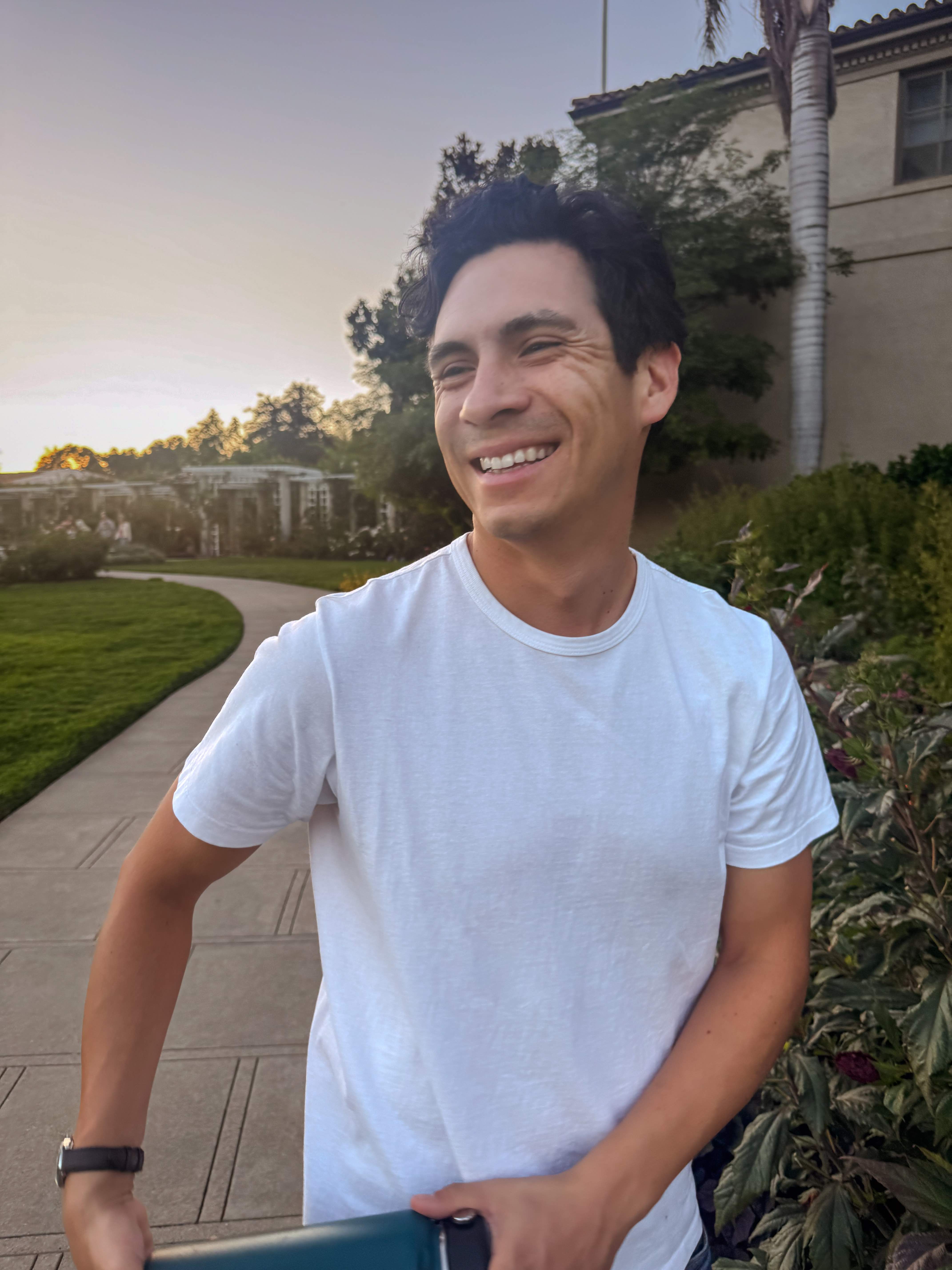 Smiling man in a white t-shirt standing on a curved path with greenery and a building in the background during sunset.