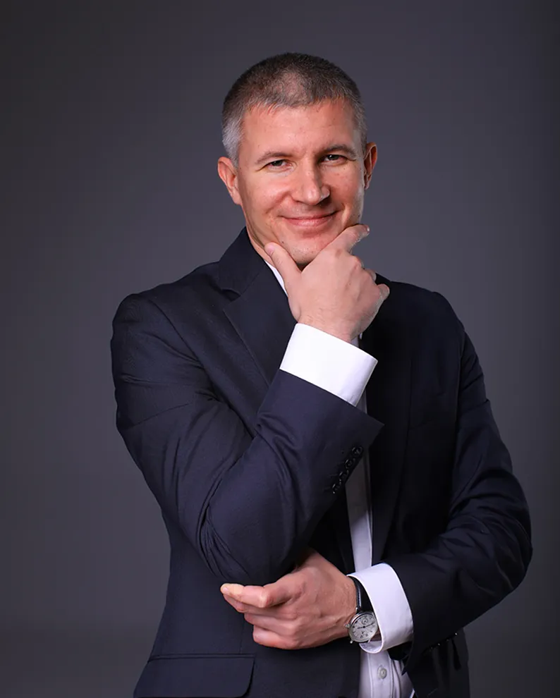 Чоловік у синьому костюмі з коротким сірим волоссям посміхається, спираючись рукою на підборіддя на сірому фоні.