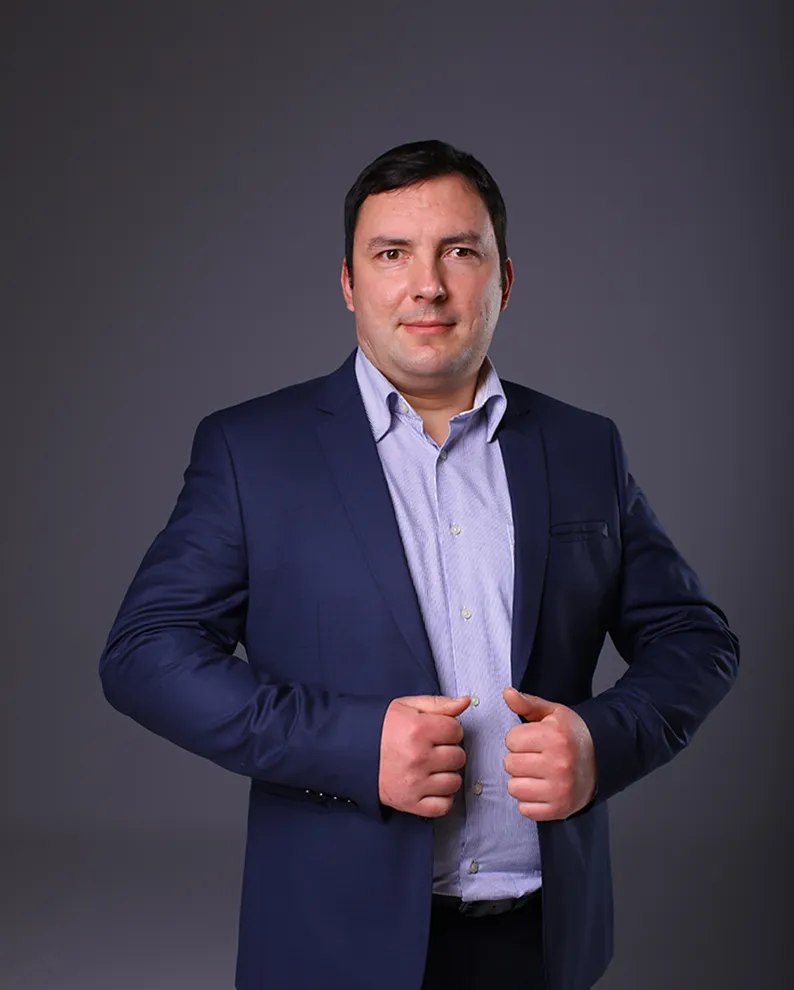 Чоловік у темно-синьому костюмі та світлій сорочці позує на сірому фоні.