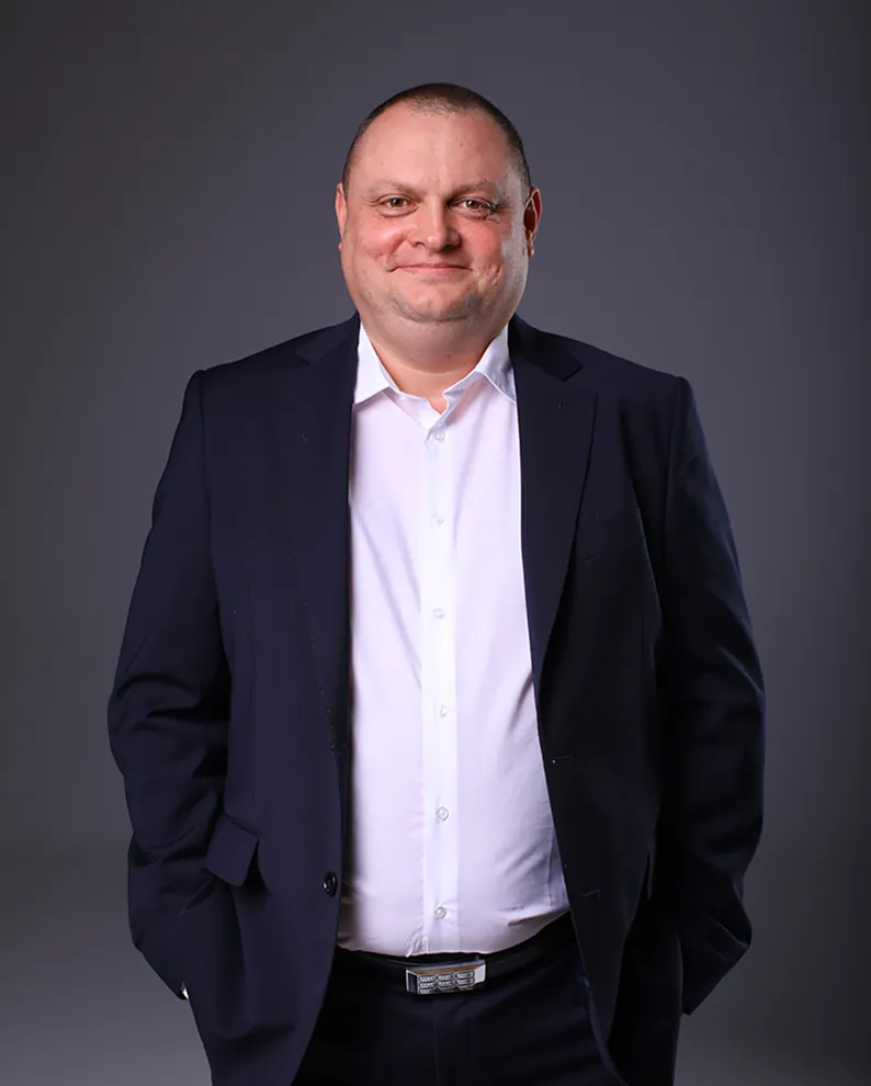 Чоловік у темно-синьому костюмі та білій сорочці посміхається на сірому фоні.