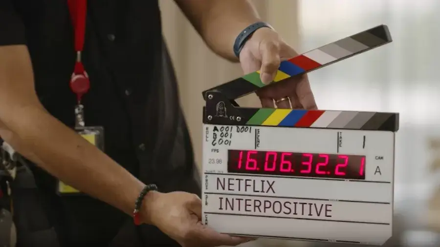 A film crew member holds a clapperboard on set displaying "Netflix InterPositive" with a digital timecode reading 16.06.32.21.