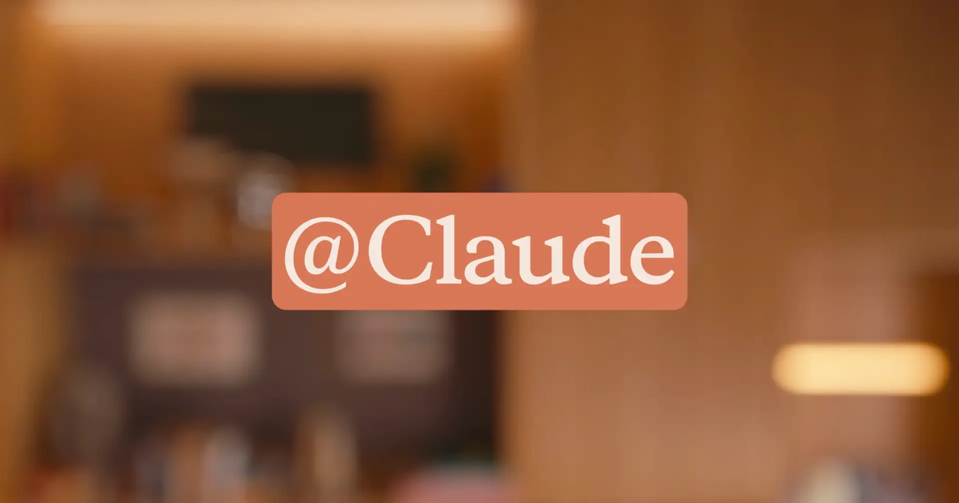 The @Claude logo in white serif text on a rounded orange badge, set against a softly blurred warm-toned room interior.