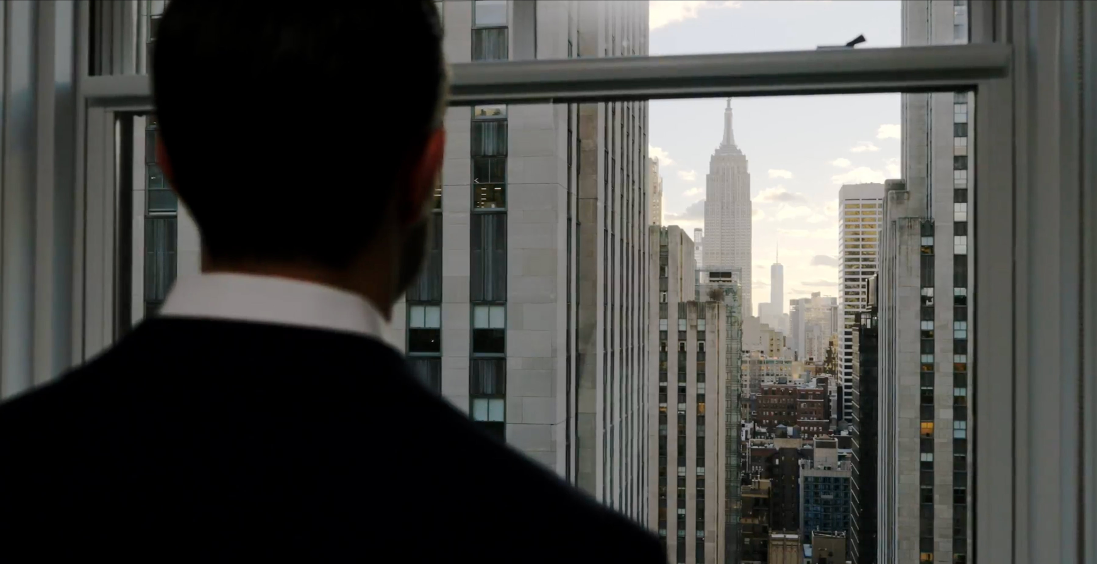 Man in suit looking out window at New York City skyline including Empire State Building.