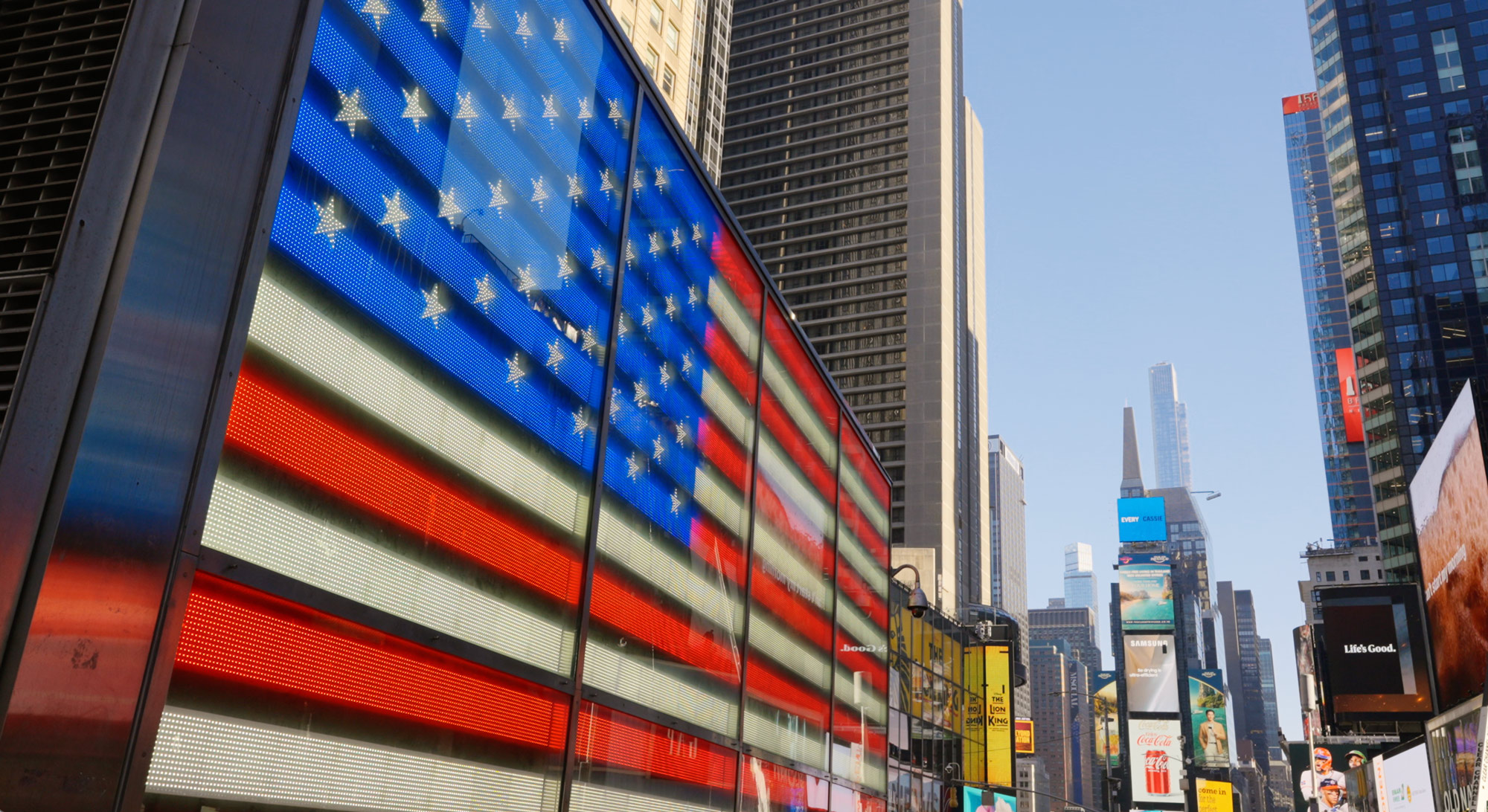 Large illuminated American flag displayed on a building in a busy city street with skyscrapers.
