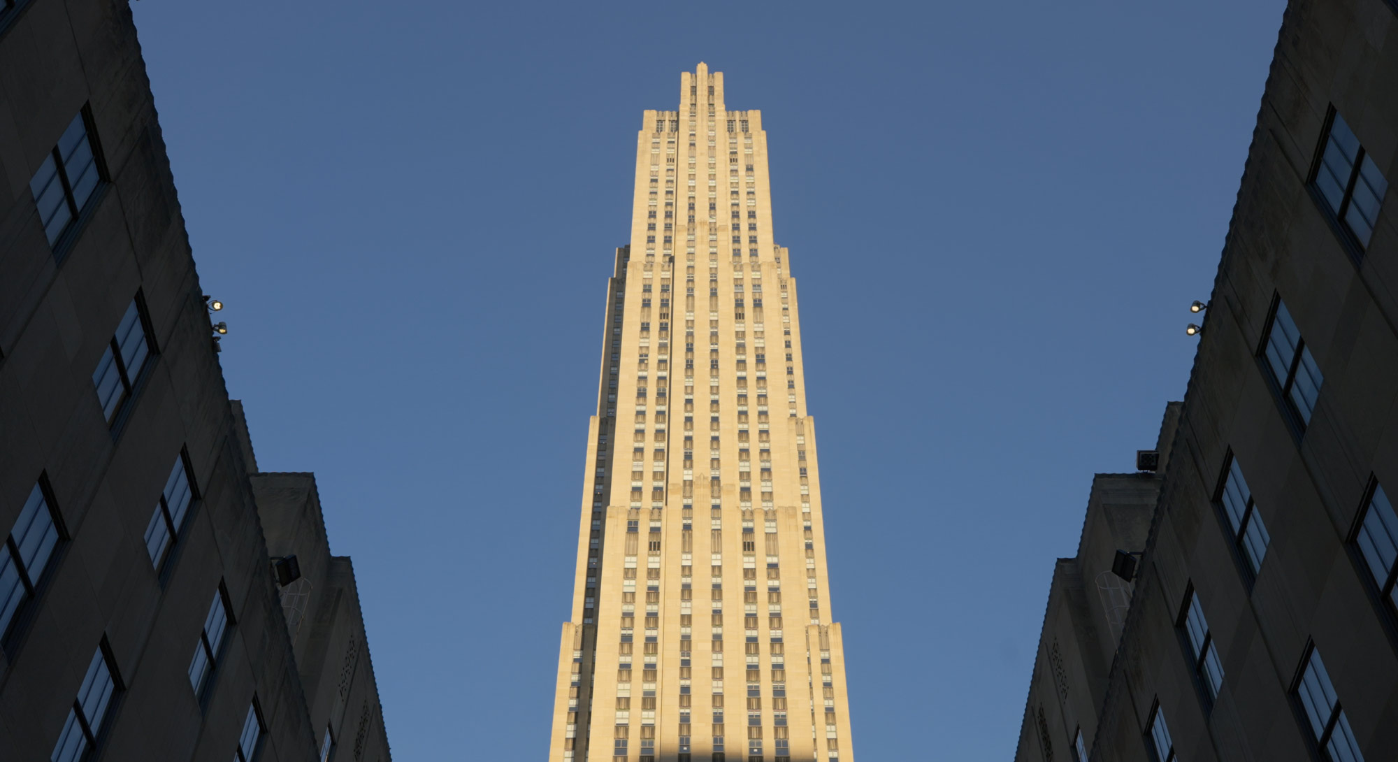 Tall beige skyscraper with vertical windows framed by two shorter buildings under blue sky.