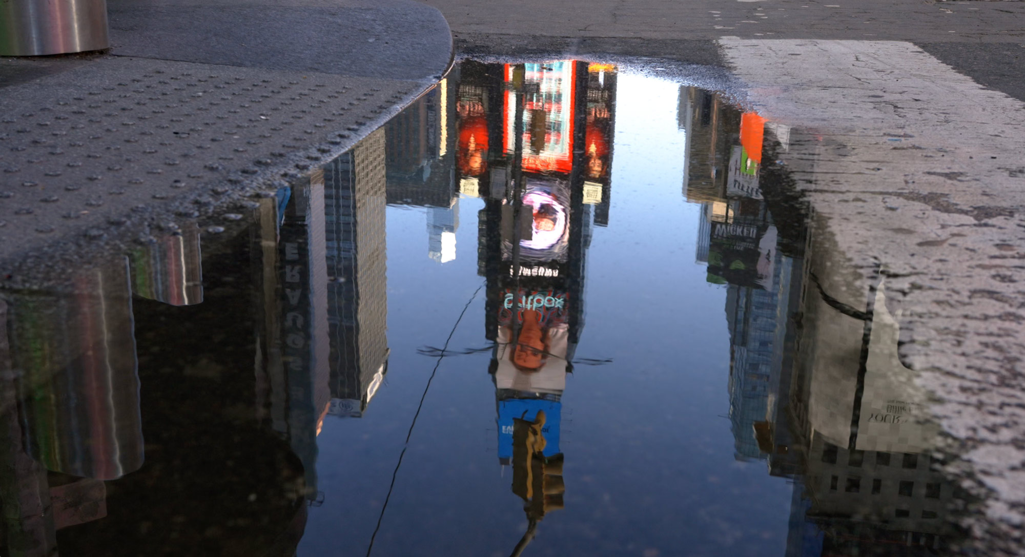 Reflection of city buildings and a billboard in a street puddle by a curb and crosswalk.