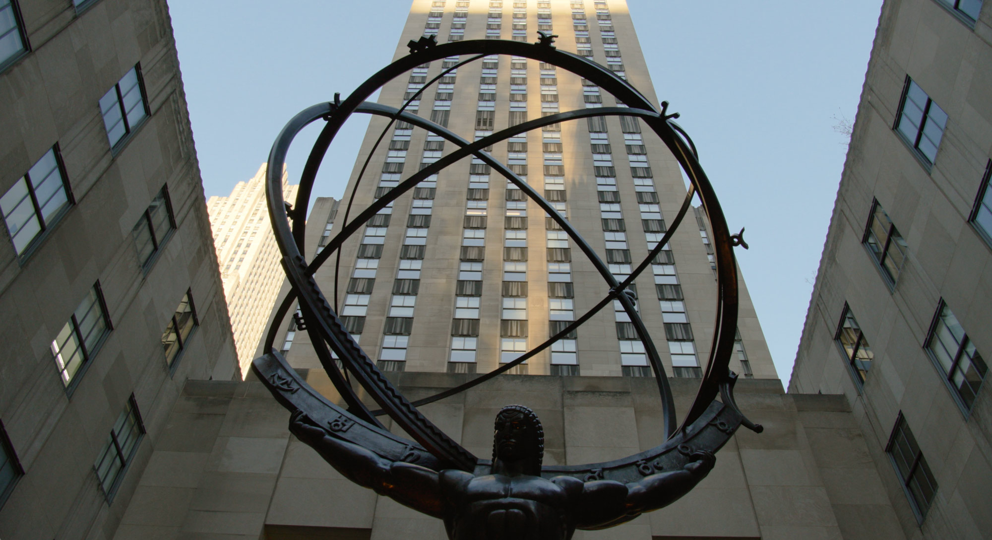 Statue of Atlas holding a celestial sphere in front of Rockefeller center under a clear sky.