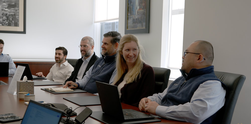 Six people seated at a conference table with laptops, engaged in a meeting in a bright office.