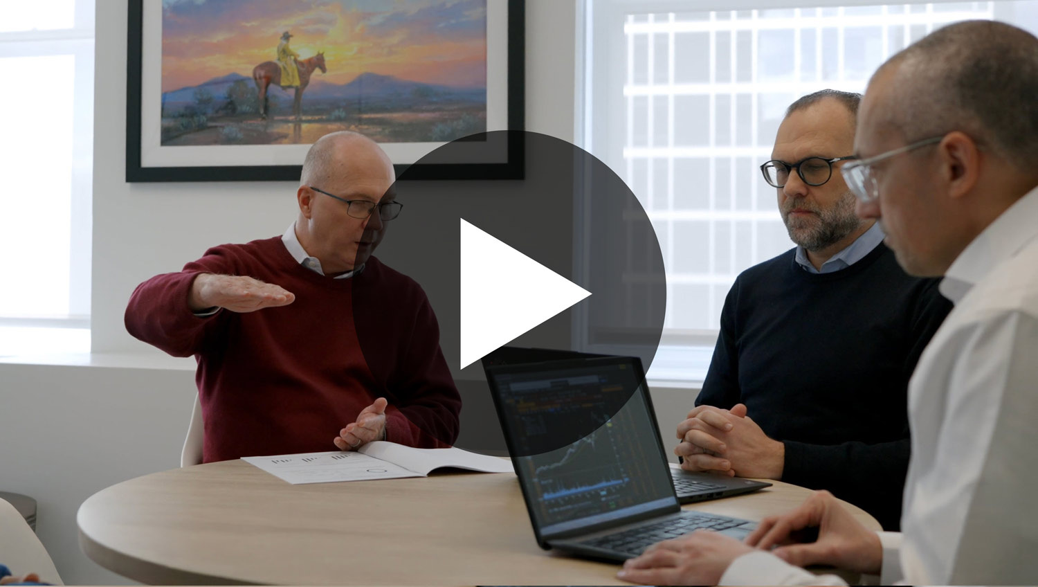 Three men in a meeting room with laptops and papers, one man gesturing while speaking.