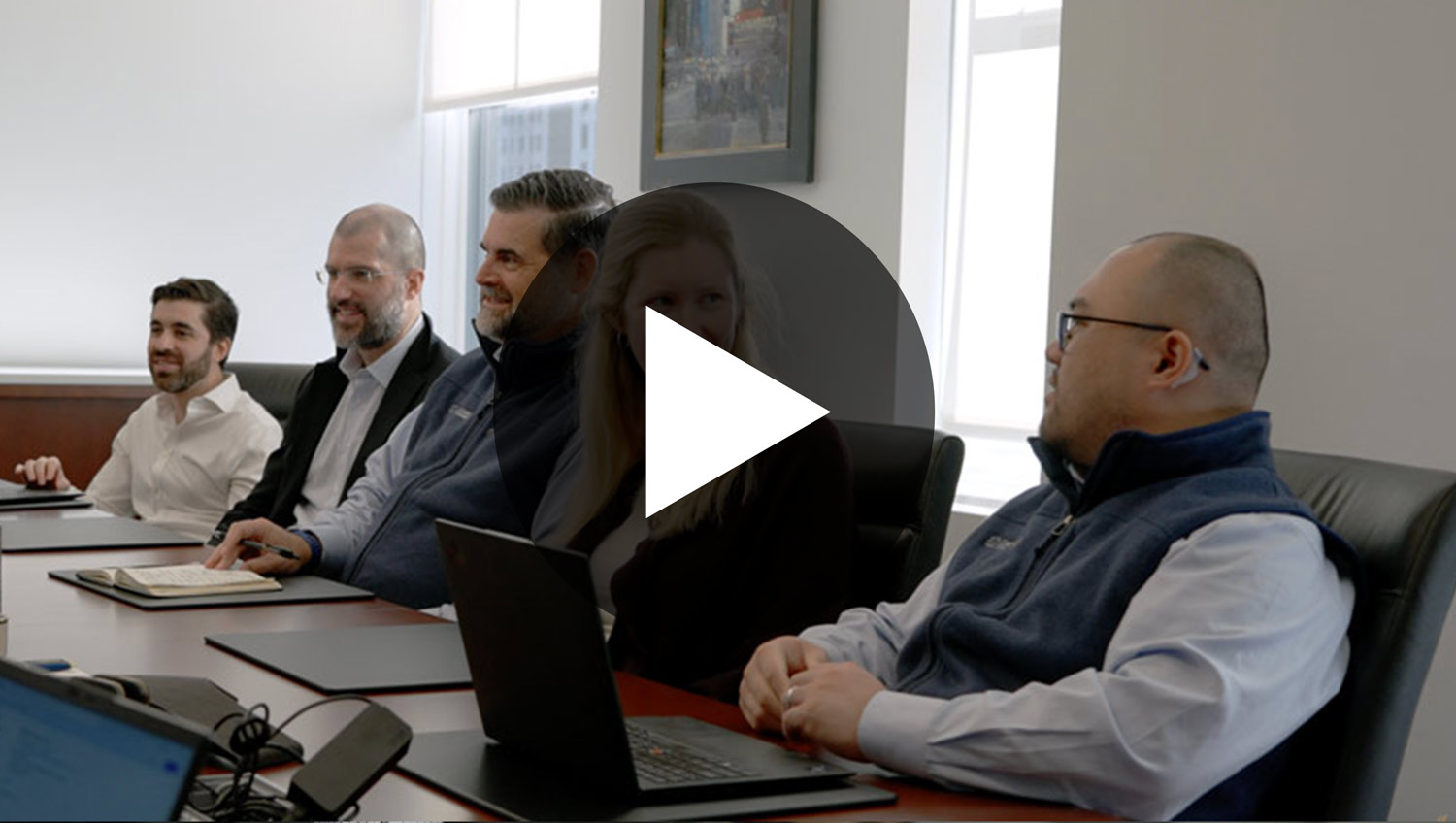 Five professionals sitting around a conference table with laptops and notebooks in a bright office.