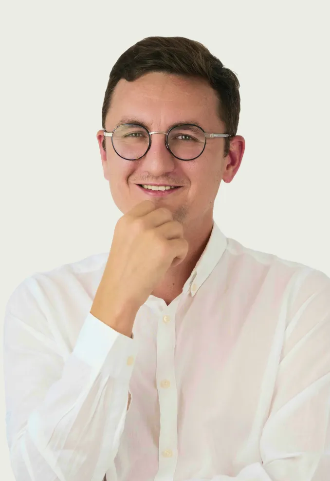 Smiling man with short dark hair and round glasses wearing a white button-up shirt resting his chin on his hand.
