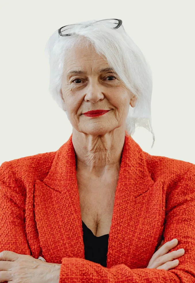 Confident elderly woman with white hair, red lipstick, and glasses on head wearing a textured red blazer with arms crossed.